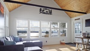 Amplia sala de estar con techo alto, ventanas grandes que ofrecen vistas al exterior, un rincón de descanso acogedor con sofá y reposapiés, y una mesa de comedor de madera.