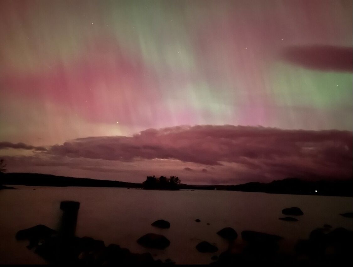 A night sky with the aurora borealis over a body of water with a silhouette of trees and rocks in the foreground.