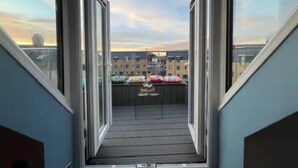 A view from a balcony with open doors, leading to a wooden deck with a railing and a small table.