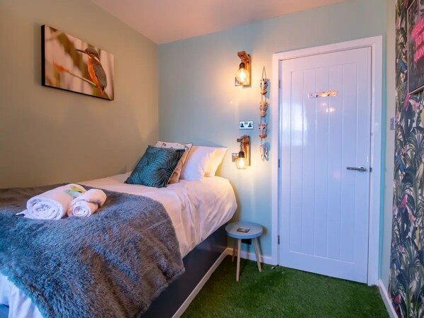 Un dormitorio con una cama, almohadas, una mesita de noche, una lámpara de pared y una puerta.