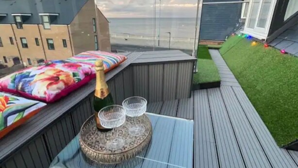 A rooftop terrace with a wooden deck, a small seating area with cushions, a bottle of champagne, and two glasses.