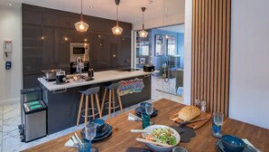 A modern kitchen with a central island, overhead pendant lights, and a dining area with a wooden table set for two.