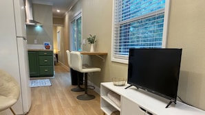 Un espacio habitable compacto con kitchenette, una zona de comedor con dos sillas, un televisor sobre un mueble blanco y una ventana con persianas.