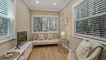 A room with a sofa, a TV, a lamp, and a chair. There are windows with blinds.