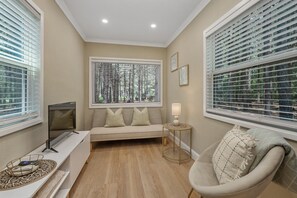 A room with a sofa, a TV, a lamp, and a chair. There are windows with blinds.