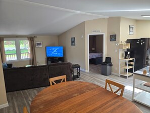 A living room with a TV, sofa, and dining area. There's a kitchen with a fridge and a coffee maker.