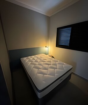 Cozy retreat with a single bed, bedside table, and lamp, plus a window with horizontal blinds.