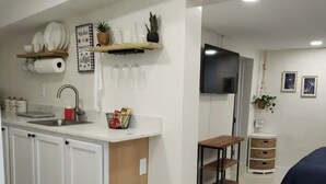 A modern kitchen with white cabinets, a wooden countertop, and a built-in sink. There is a small dining area with a table and chairs, and a bed in the background.
