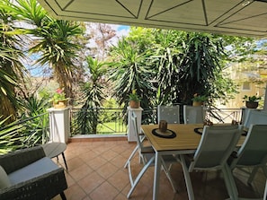 A covered patio with a wooden table and chairs, surrounded by potted plants and a view of greenery.