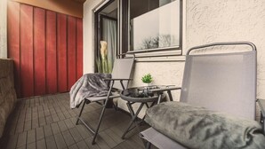 A small balcony with a red wooden wall, a window, two chairs, a small table, and a potted plant.