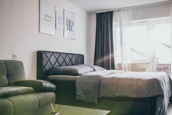 A modern hotel room with a bed, a green armchair, and a view of the city through large windows.