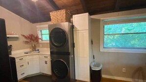 A compact kitchen with a washer and dryer, wooden cabinets, a window with a view of greenery, and a mop leaning against the wall.