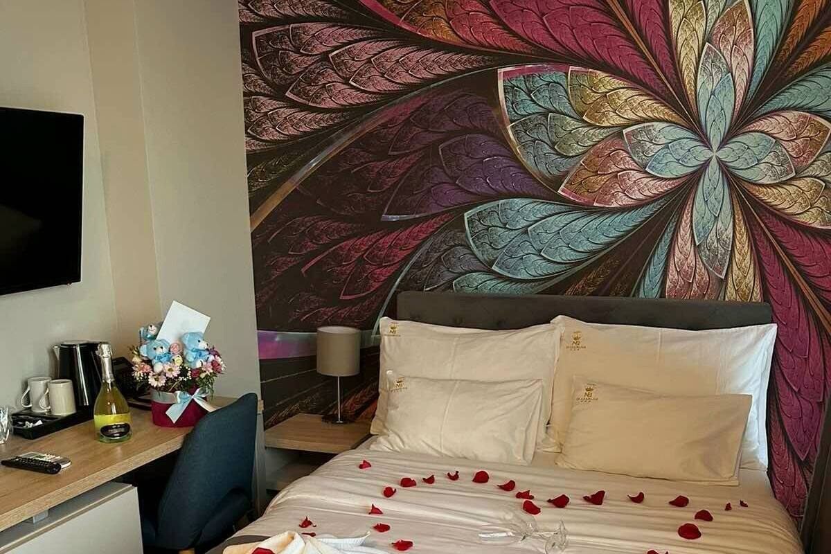 A hotel room with a bed adorned with rose petals and towels, a desk with a bottle of wine, a vase of flowers, and a large decorative wall panel.