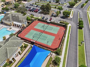Een tennisbaan omringd door een zwembad en een overdekt terras, met een parkeerplaats en woongebouwen op de achtergrond.