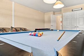 A pool table with balls and a cue stick, a sofa with cushions, and a door in a room with beige walls.