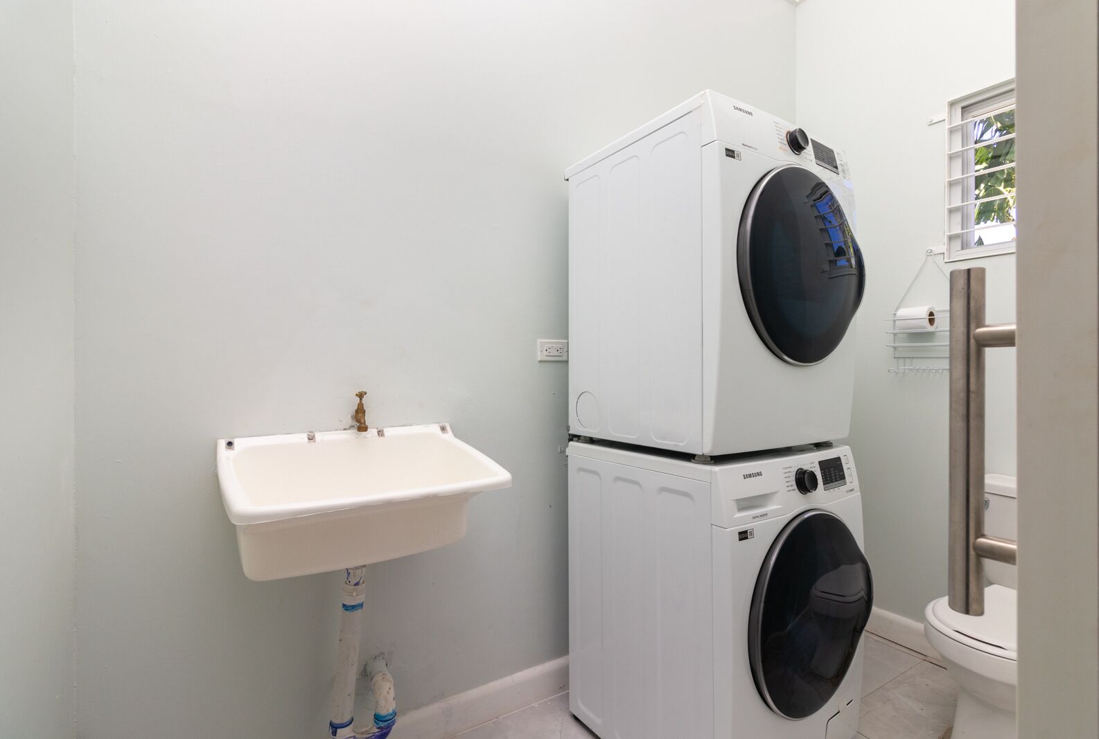 Uma lavanderia compacta com máquina de lavar e secadora empilhadas, ambas brancas, uma pia branca com torneira de latão e uma janela com vista para árvores.