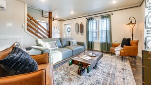 A modern living room with a sofa, coffee table, and a chair. There is a staircase with wooden railings and a view of the outside through a window with curtains. - Historic Hollister Home: Shop, Dine & Drink Nearby (Hollister)