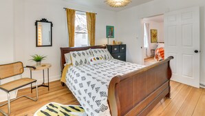 Ein Schlafzimmer mit einem hölzernen Bett, einem kleinen Tisch mit Pflanze, einem Stuhl und Blick in einen anderen Raum durch eine geöffnete Tür.