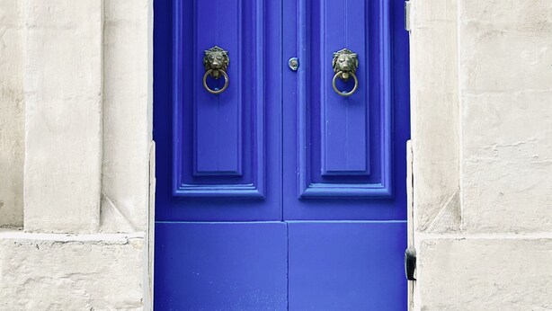 Une porte bleue, surmontée d’un élément décoratif en métal et d’une plaque portant un numéro.