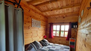 A wooden cabin bedroom with a bed, curtains, a window with red curtains, and a wooden ceiling.