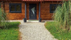 A wooden cabin with a gravel path leading to a dark door, surrounded by greenery.