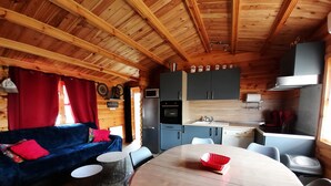 A cozy cabin interior with a wooden ceiling, a blue sofa, a round dining table, and a kitchen area with cabinets and appliances.