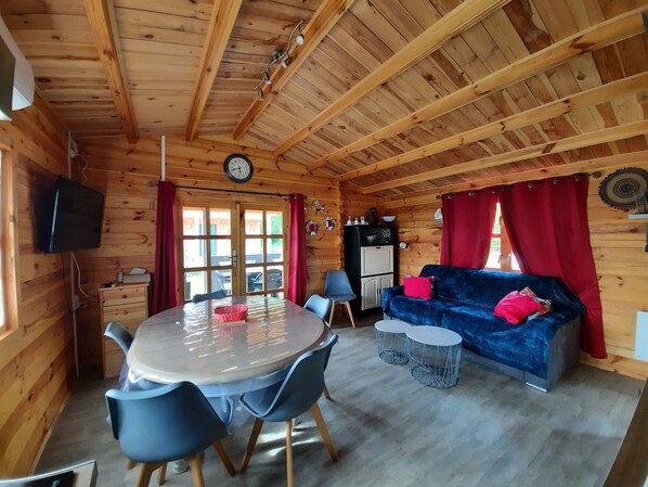 Interior de una cabaña de madera con una mesa de comedor, sillas, un sofá y un televisor.
