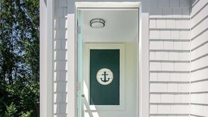 Un bâtiment blanc doté d’une terrasse en bois menant à une porte avec un cadre vert et ornée d’une ancre.
