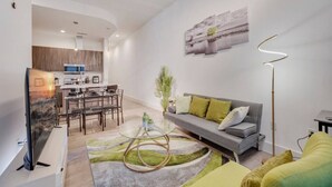 Una sala de estar moderna con un sofá gris, una mesa de centro de vidrio y una zona de comedor, con una cocina al fondo.