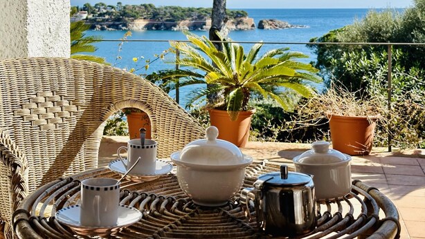 A wicker chair and table set with white teacups, a teapot, and a sugar bowl on a balcony overlooking a coastal view. - Maset CostaBravaSi - with pool, views, parking (Sant Feliu de Guíxols)