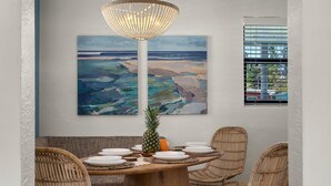 A dining area with a wooden table set for four, surrounded by wicker chairs, a pineapple centerpiece, and a large abstract painting on the wall.