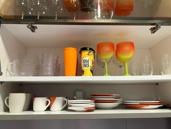 A shelf with various glassware and dishes, including clear glasses, orange and green tumblers, and white and orange plates. - Makia Beach Resort Experience Exclusive (Ipojuca)