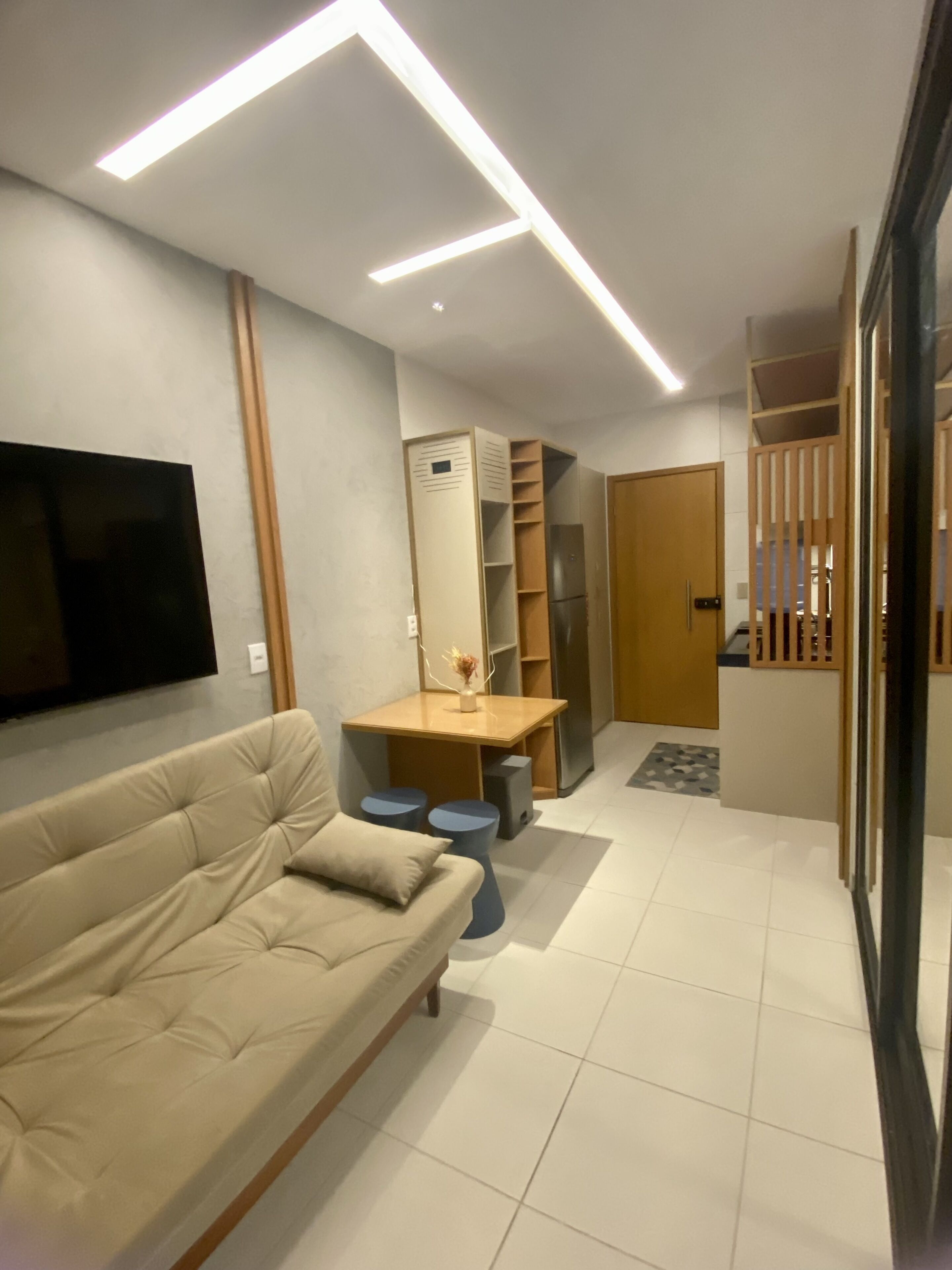 Una sala de estar moderna con un sofá color beige, un televisor de pantalla plana montado en la pared, una mesa de centro de madera y un mueble empotrado con refrigerador.
