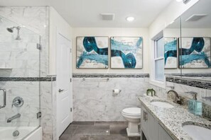 Un baño moderno con ducha de mármol, inodoro, lavamanos y un espejo grande. Hay pinturas abstractas en la pared sobre el espejo.
