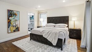 Un dormitorio con cama, mesita de noche, lámpara, un cuadro y una ventana.