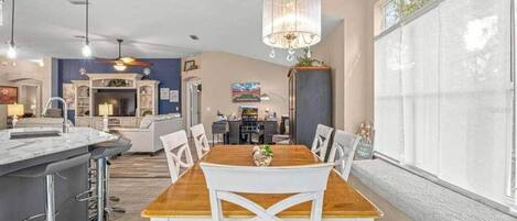 A modern kitchen with a dining area, featuring a wooden table, white chairs, and a chandelier.