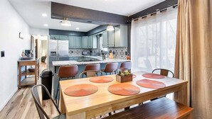 A modern kitchen with a central island, a dining area with a wooden table and chairs, and a view of the city through large windows. - The Den is a cozy and welcoming 3 bed, 2 bath home with zero entry design (Wichita)