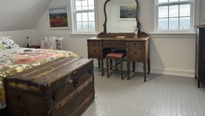 A bedroom with a wooden dresser, a bed with floral bedding, a small chair, and a large window with a view of the sky. - Charming rustic retreat with Bay of Fundy Views, escape to our serene haven. (Hampton)
