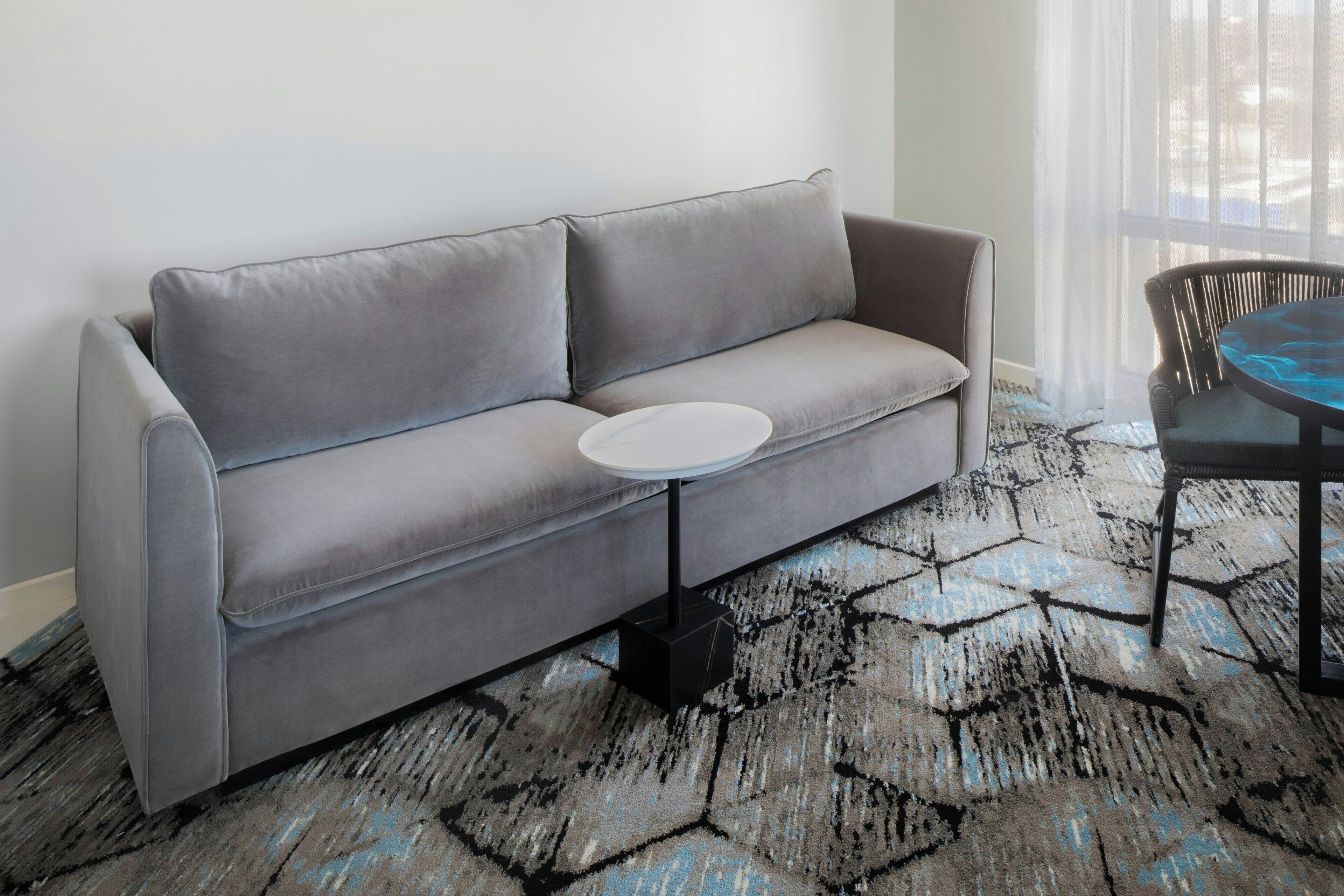 Ein graues Sofa mit einem runden weißen Beistelltisch, einem gemusterten Teppich und einem Sessel mit blauem Kissen.