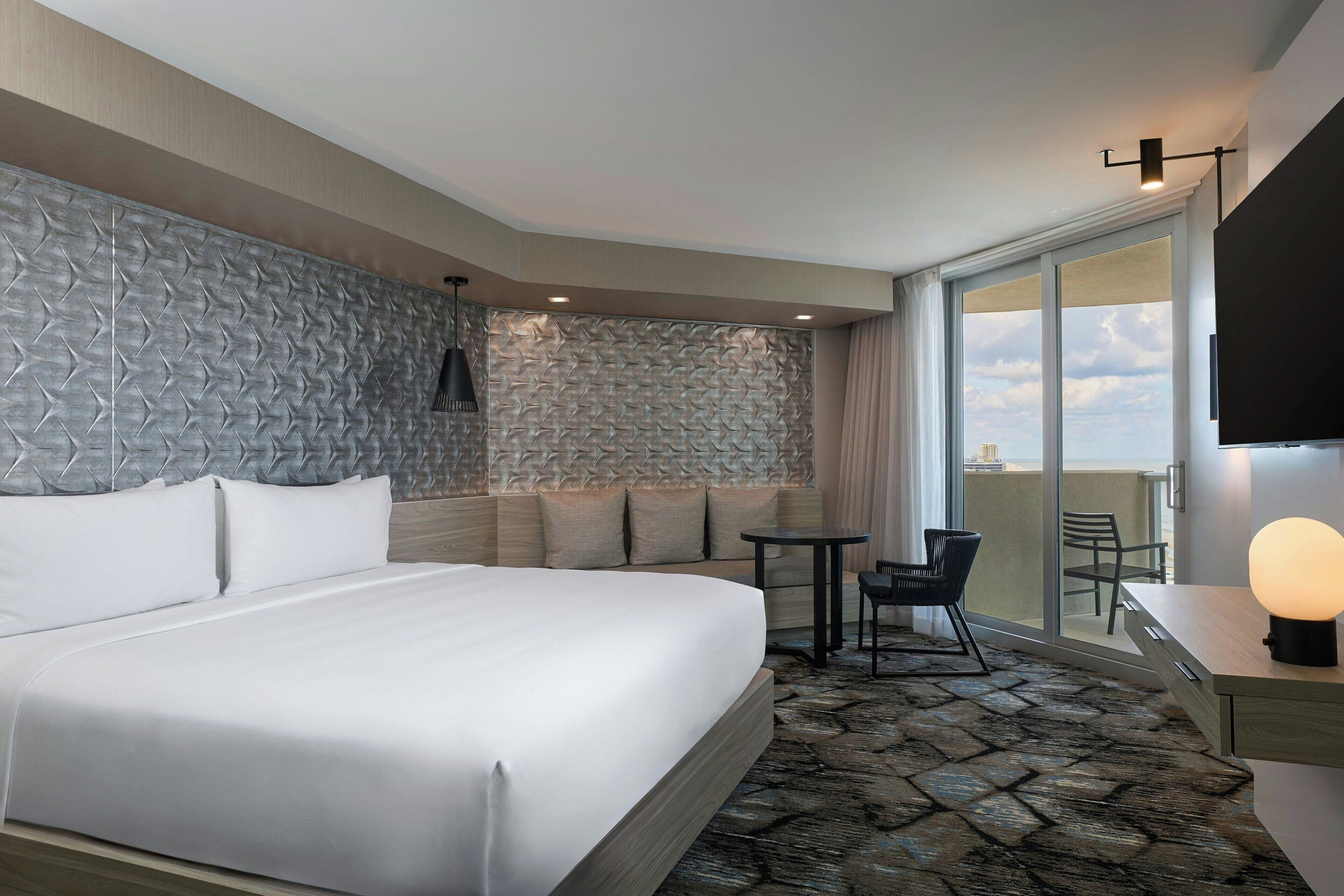 A hotel room with a large bed, a stone-patterned floor, a seating area with a table and chairs, and a balcony with a view.