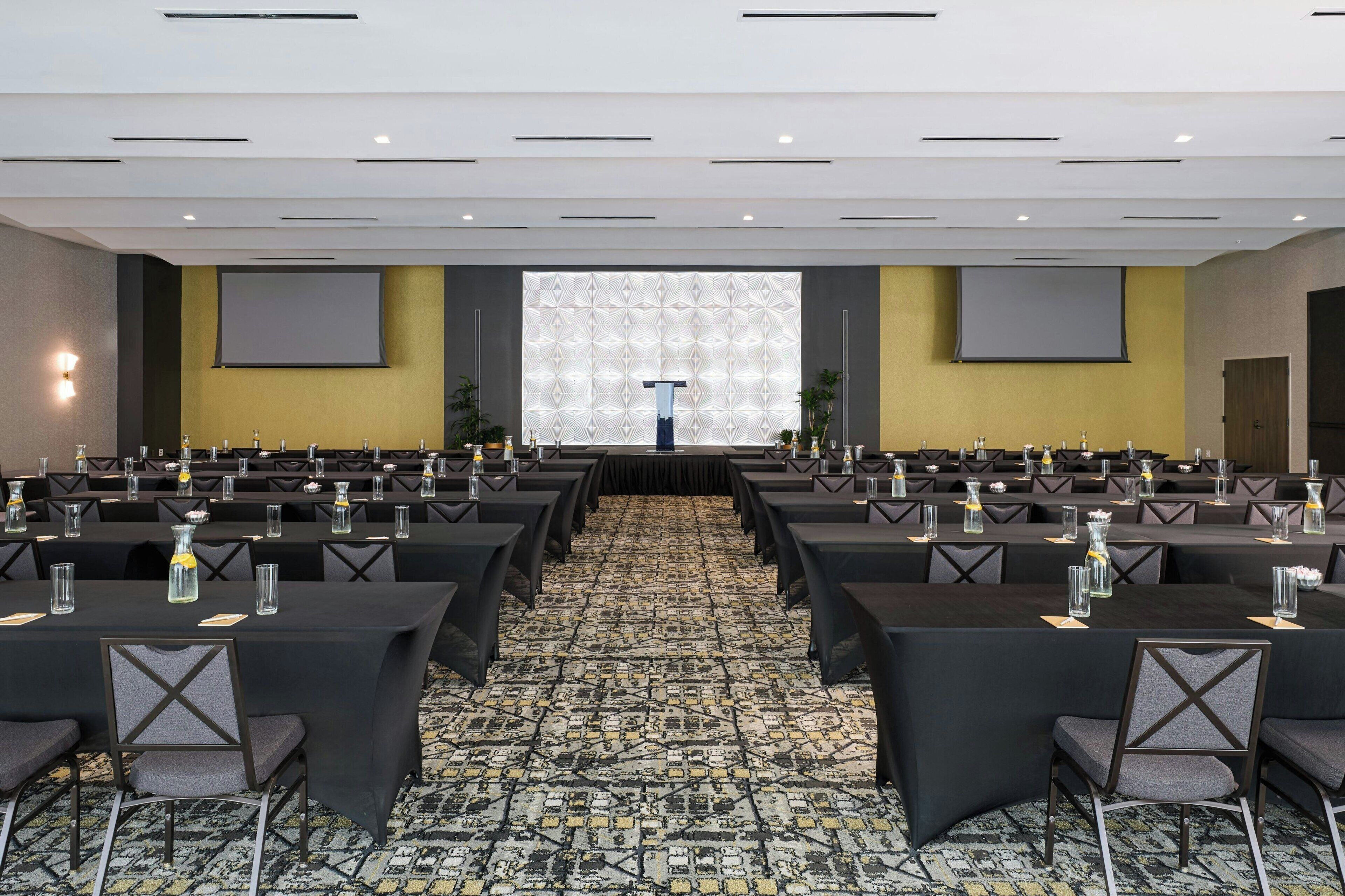 Un salón de conferencias con varias mesas y sillas, una pantalla de proyector y un atril.