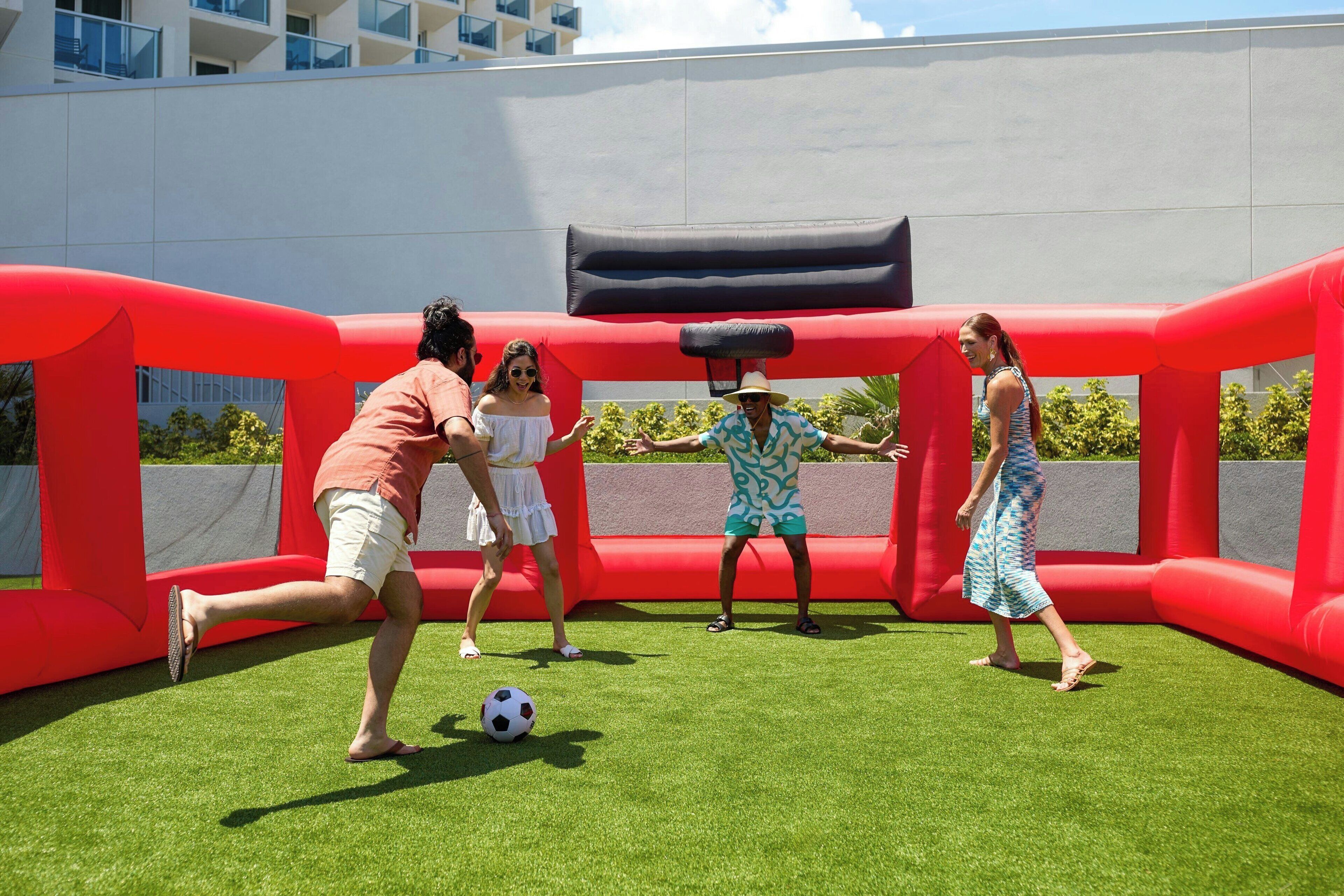 Un grupo de personas jugando fútbol en un campo cubierto de césped con un obstáculo inflable rojo.