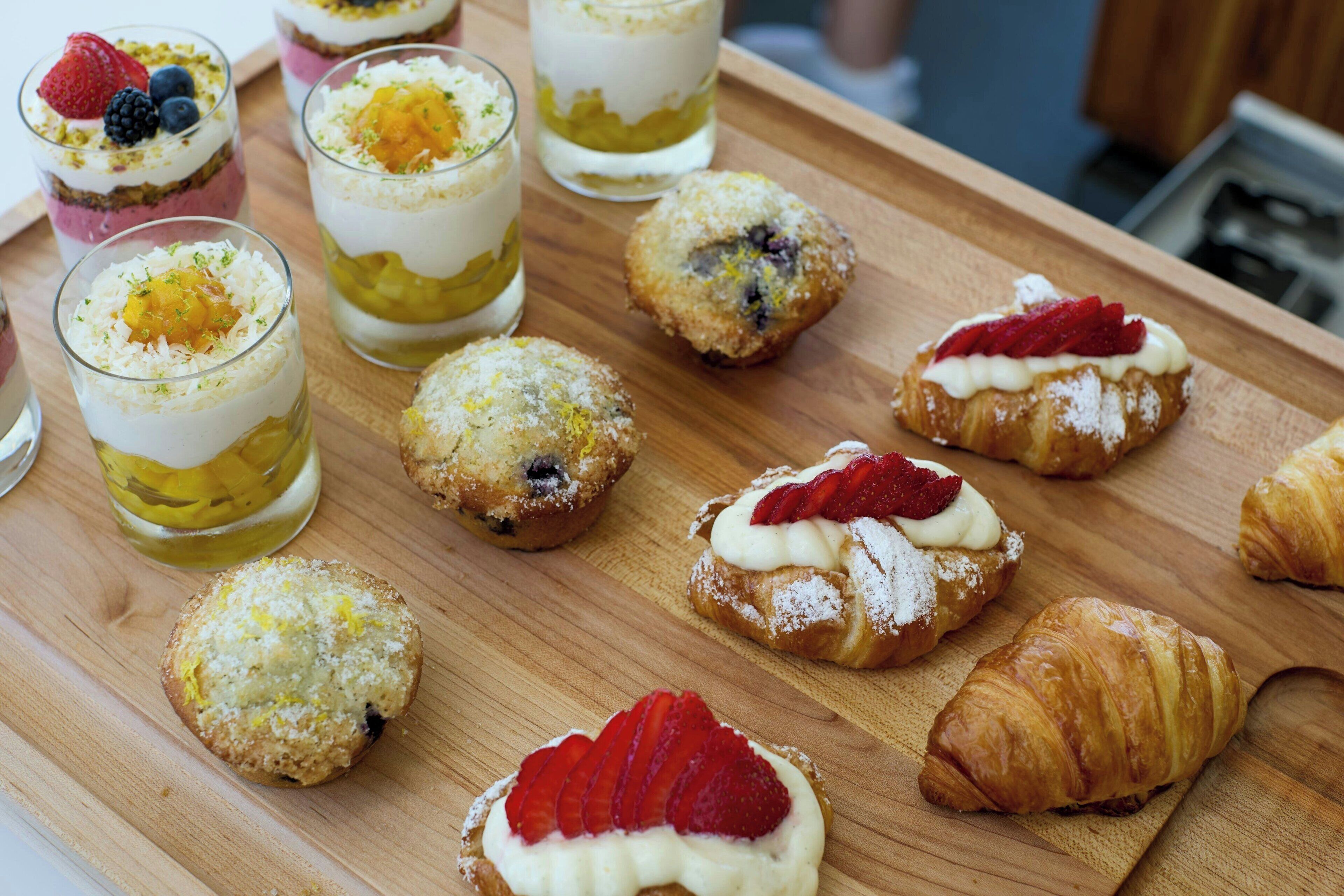 A wooden tray with various pastries and desserts, including croissants and muffins topped with fruits and cream.