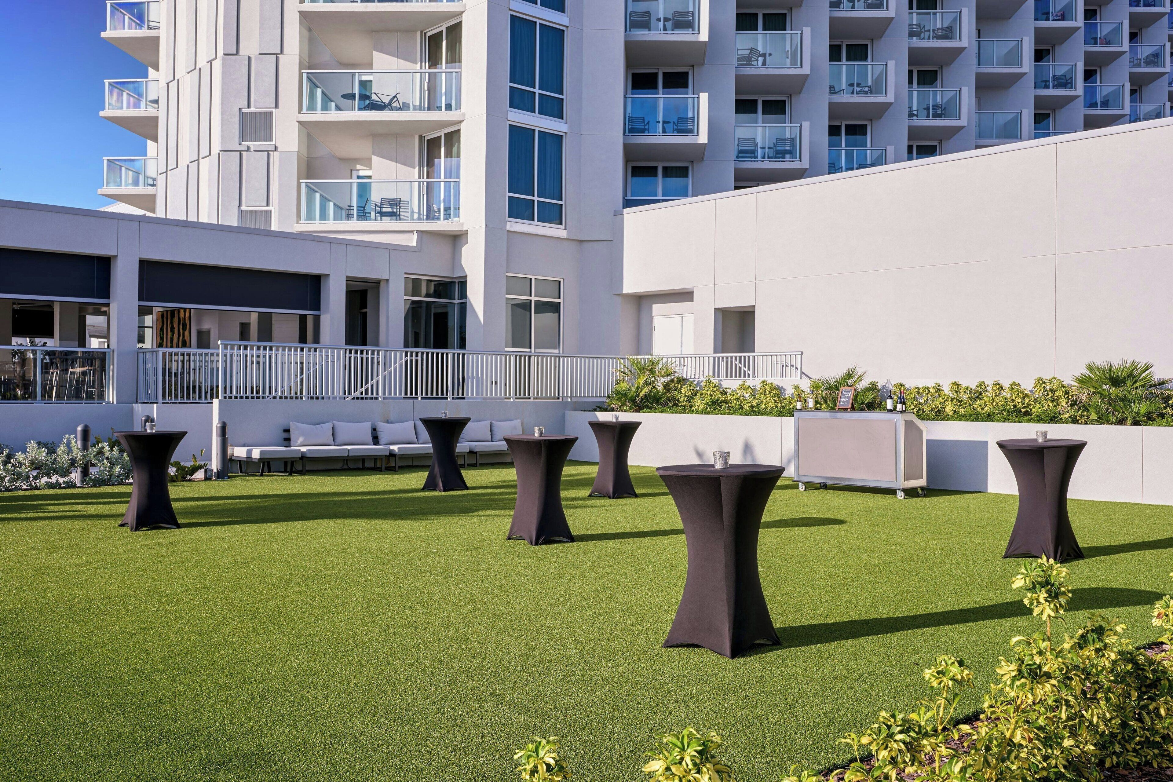 Una terraza en la azotea con césped artificial, varias mesas redondas y un edificio blanco con balcones al fondo.