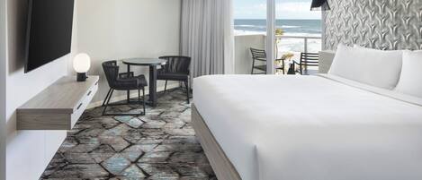 Quarto de hotel com uma cama grande, uma televisão fixada na parede, uma mesa pequena com cadeiras e vista para o oceano por grandes janelas.