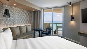 Et hotelværelse med en stor seng, en sofa, et lille bord og en stol. Der er en balkon med udsigt over havet.