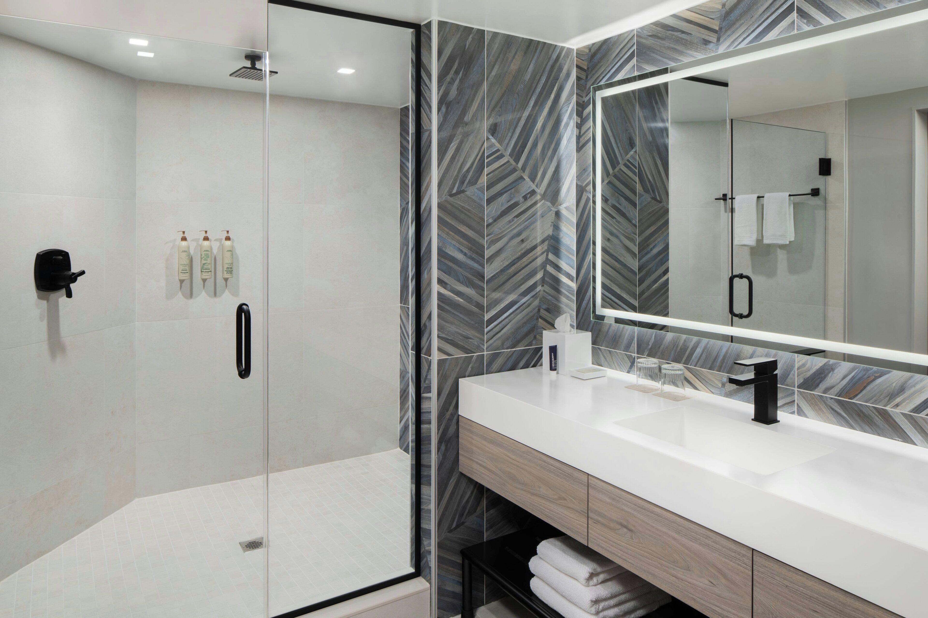 A modern bathroom with a glass-enclosed shower, a white sink with a black faucet, and a large mirror with a herringbone-patterned backsplash.