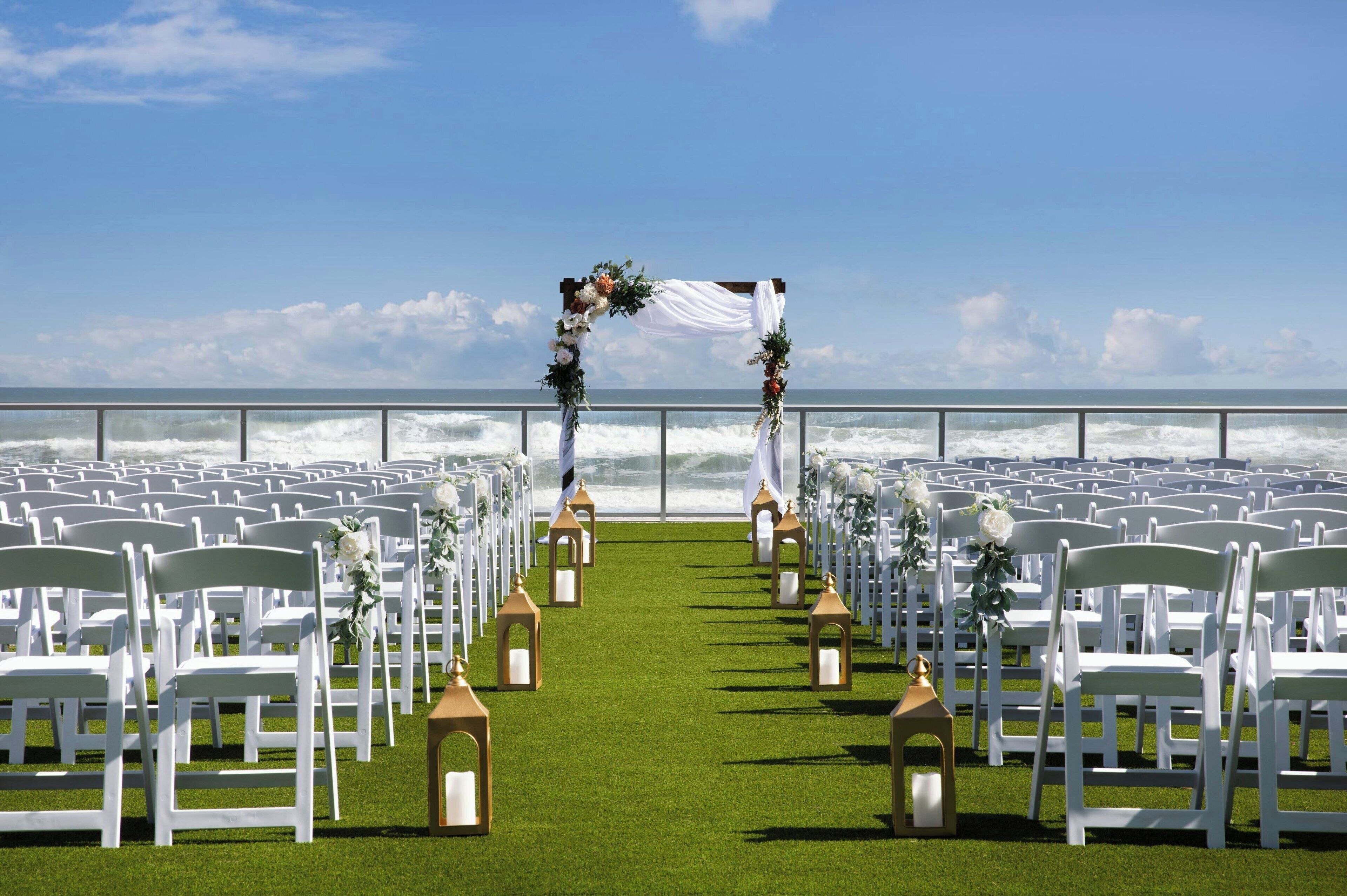 A beachside ceremony aisle with white chairs and lanterns, leading to a wooden arch decorated with flowers.