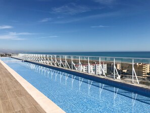 A long swimming pool with a glass railing, overlooking a beach and buildings. - Global Vacacional: Modern flat with swimming pool and terrace on the beach of Canet (Canet d´en berenguer)