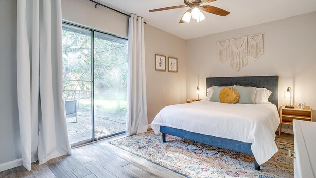 A bedroom with a large bed, a ceiling fan, a view through a sliding glass door, and a macramé wall hanging.